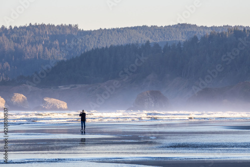 Person photographing ocean waves on a wide Oregon beach with offshore sea stacks, mist, and forested coastal hills