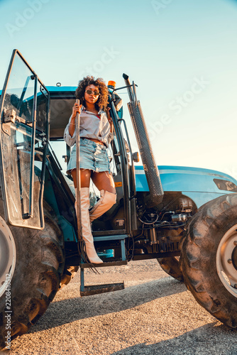 Woman in sunglasses stands on tractor steps near large farm equipment by field edge, wearing striped shirt, denim skirt, and tall boots with bright daylight and open rural sky.