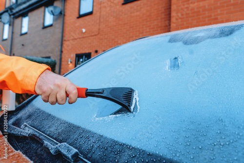 Person scraping ice off a car windshield in a residential area during winter morning