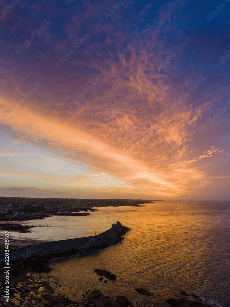 Fototapeta premium Aerial view of coastal pier at golden sunset. Warm light reflects on water and stone breakwater. Calm sea meets land with sandy beach nearby. Spiddal, Ireland, Galway bay. Travel and tourism.