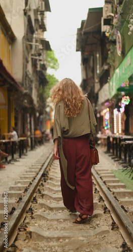 A Happy Young Woman with Curly Hair Walks and Smiles on the Iconic Train Street in Hanoi, Vietnam, Experiencing the Unique Urban Atmosphere and Local Culture.