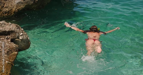 A Young Woman in a Red Bikini Snorkels in Crystal Clear Turquoise Sea Water Near a Rocky Coastline on a Beautiful Sunny Summer Day, Enjoying Her Vacation.