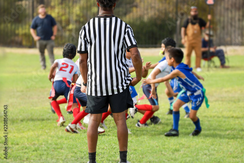 A football referee in a black and white striped uniform stands on a grass field, watching young children play an active game of youth flag football.