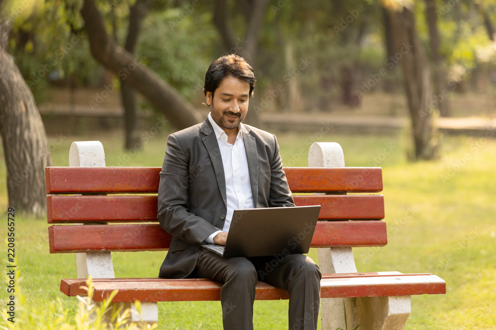 Fototapeta premium Indian Young businessman is sitting at the park and using laptop. Businessman using laptop outdoor