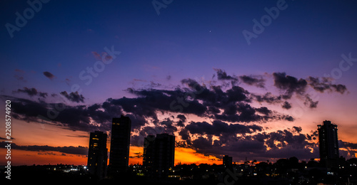 A breathtaking city skyline silhouette under a dramatic twilight sky filled with deep purple clouds and a vibrant orange sunset glow, featuring a single bright star or planet visible