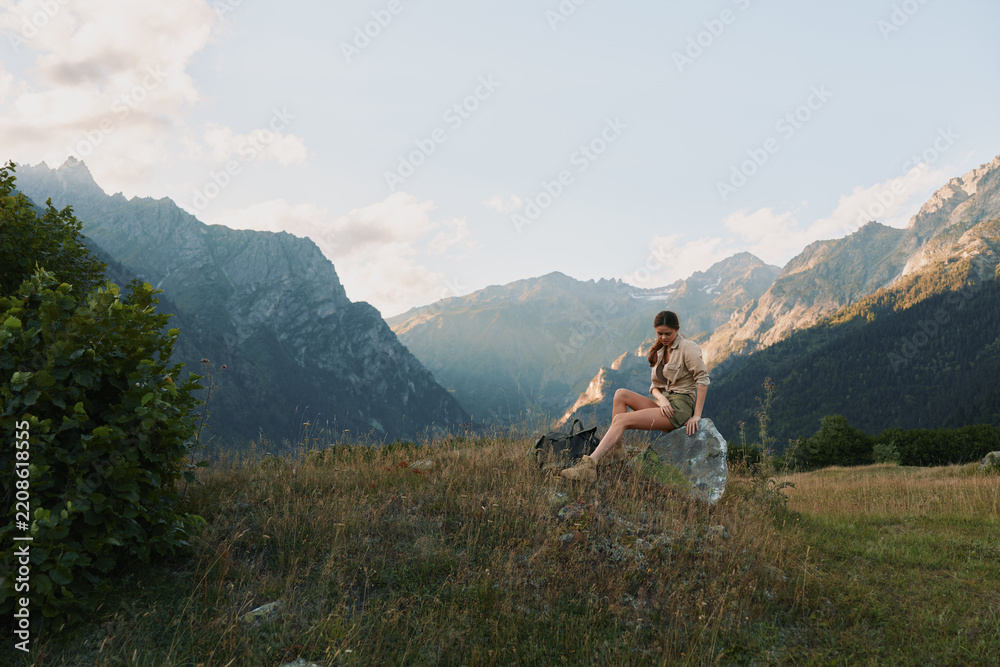 Obraz premium Joyful woman outdoors, seated on a meadow with mountain backdrop, warm sunlight, casual attire, relaxed posture, tranquil nature scene, gentle breeze, vibrant colors, lifestyle moment