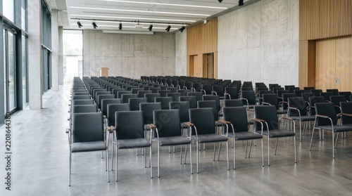 A large, empty conference hall is filled with rows of black chairs arranged in a lecture setting. The hall features a concrete wall, wooden paneling, and a raised stage.