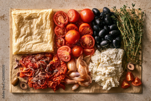 Mediterranean ingredient flat lay with pastry, tomato, olive, feta, garlic, thyme, anchovy and sundried tomato arranged for gourmet savoury cooking.