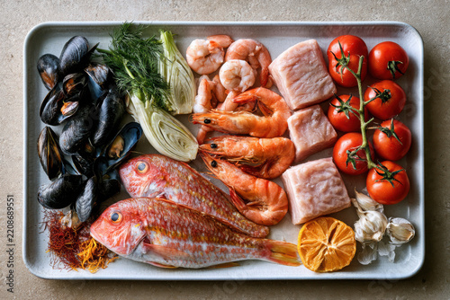 Mediterranean seafood flat lay with whole fish, mussel, prawn, fennel, tomato, garlic, citrus and saffron arranged on a tray for gourmet cooking.