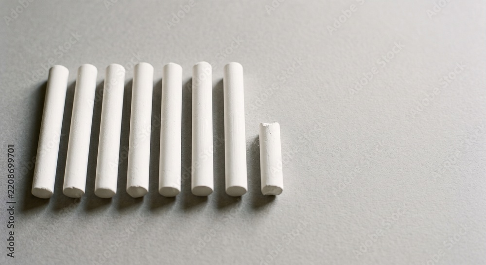 Fototapeta premium Row of blank white chalk sticks with one short broken piece on a light grey paper background with copy space