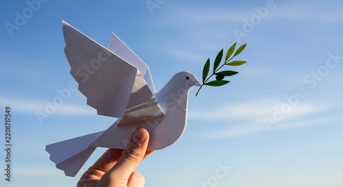 Gentle hand holding paper dove with fresh olive branch beneath blue sky representing peace harmony hope and goodwill to all