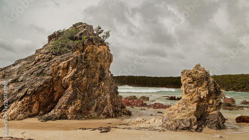 Rocky, rugged rock formations, remnants after strong weathering and erosion, stand out amongst smaller rocks under an overcast sky on a windy day at Pambula Beach inlet in New South Wales, Australia.