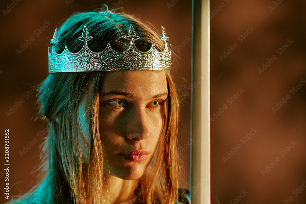 Obraz premium Young adult Caucasian woman wearing silver crown holding sword, looking away with serious expression. Concept representing power, leadership, suitable for fantasy gaming and storytelling