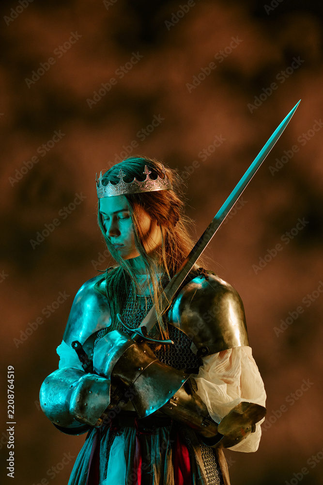 Obraz premium Vertical image of Caucasian young adult woman wearing medieval knight armor with crown holding sword looking down. Female warrior concept useful for fantasy game promotion, vertical