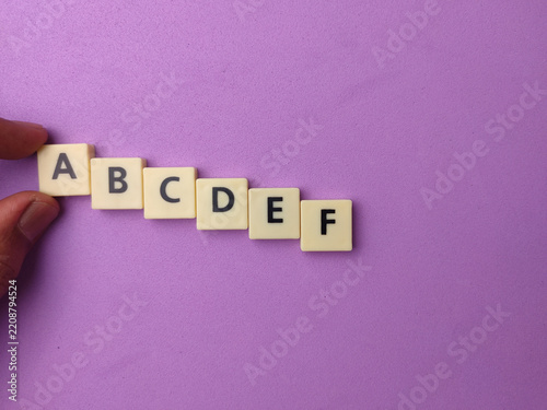 A hand holds letter tiles spelling the alphabet sequence A to F on a purple background
