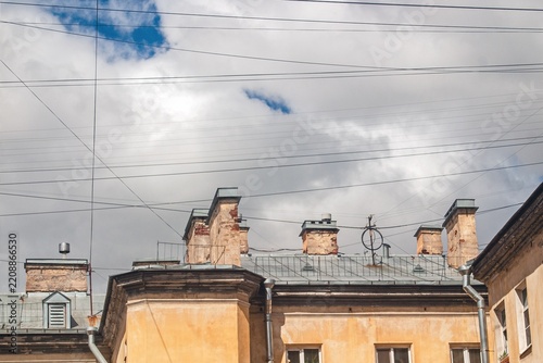 Weathered building with chimney stacks captures the interplay of rooftops and tangled wires against a backdrop of clouds and blue sky.