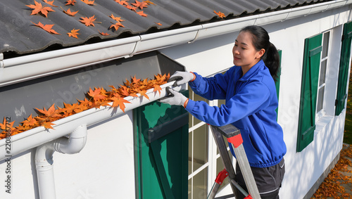 Woman in protective gloves clears fallen autumn leaves from a house gutter while standing on a ladder beside green shutters and a white exterior wall