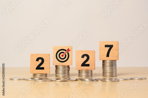 Financial growth and investment goal concept for 2027. Wooden blocks with 2027 and target icon placed on stacked coins, representing economic planning, profit strategy, savings and business success.