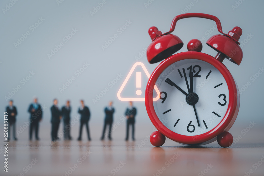 Fototapeta premium Red alarm clock with glowing warning sign and miniature business people symbolize urgent deadlines from time pressure, project delays and business risk awareness.