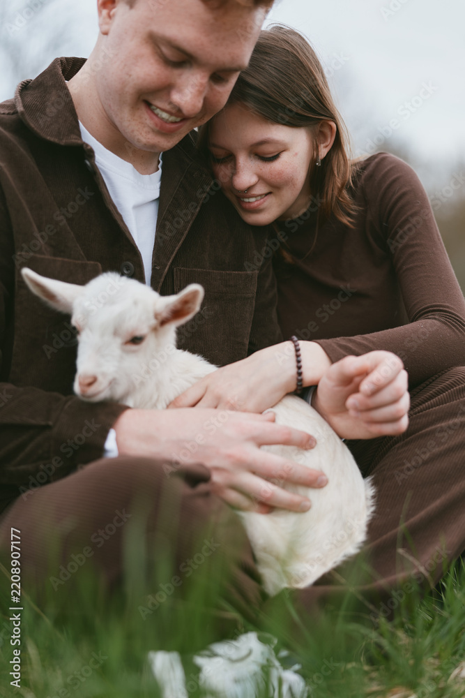 Obraz premium Cheerful couple outdoors with a white goat kid, warm clothes, genuine smiles, sunlit grass meadow, relaxed pose, candid lifestyle moment, natural colors, rustic vibe, playful affection