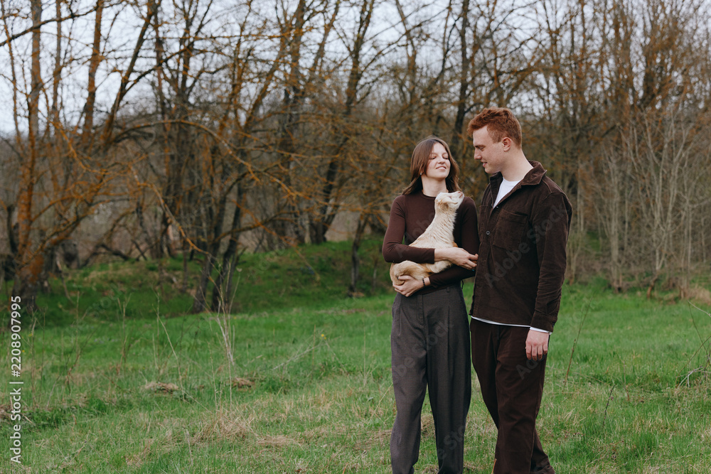 Obraz premium A couple outdoors with a goat kid in a grassy field, smiling at each other. A warm, candid lifestyle moment in a natural setting, perfect for family, animals, and nature themed storytelling.