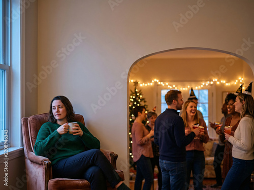 Woman sitting alone and resting in armchair during a holiday party. She feels pensive while others celebrate.
