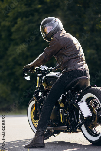 Rider in leather jacket and helmet posing on a custom vintage motorbike outdoors, enjoying the open road and freedom