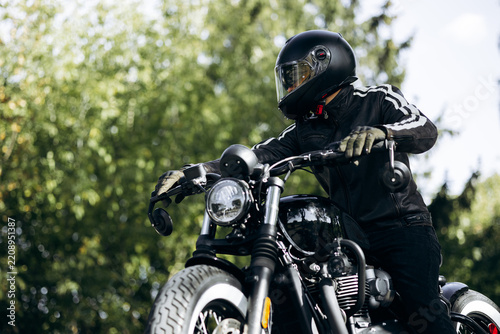 Biker wearing full-face helmet and leather jacket, riding a black vintage custom motorcycle on a road with green trees