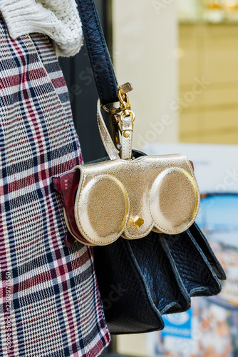 Gros plan d’un accessoire doré fixé à la bandoulière d’un sac à main noir, porté avec une veste à carreaux.
