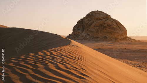 Desert landscape at golden hour with a large sand dune in the foreground and a solitary butte rising from the flat arid plain behind, distant mountains on the hazy horizon