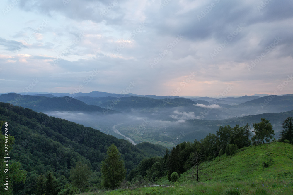 Fototapeta premium Scenic drone panorama of winding mountain river in green valley surrounded by forested slopes and morning fog