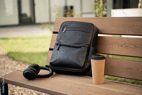 Black backpack, wireless headphones and takeaway coffee arranged on a wooden bench outdoors, representing mobile work, commuting, travel and modern urban lifestyle.