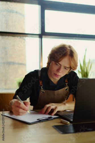 Vertical image of Caucasian young small business owner writing notes while working on laptop. Ideal for small business management, entrepreneurship, and cafe accounting concepts