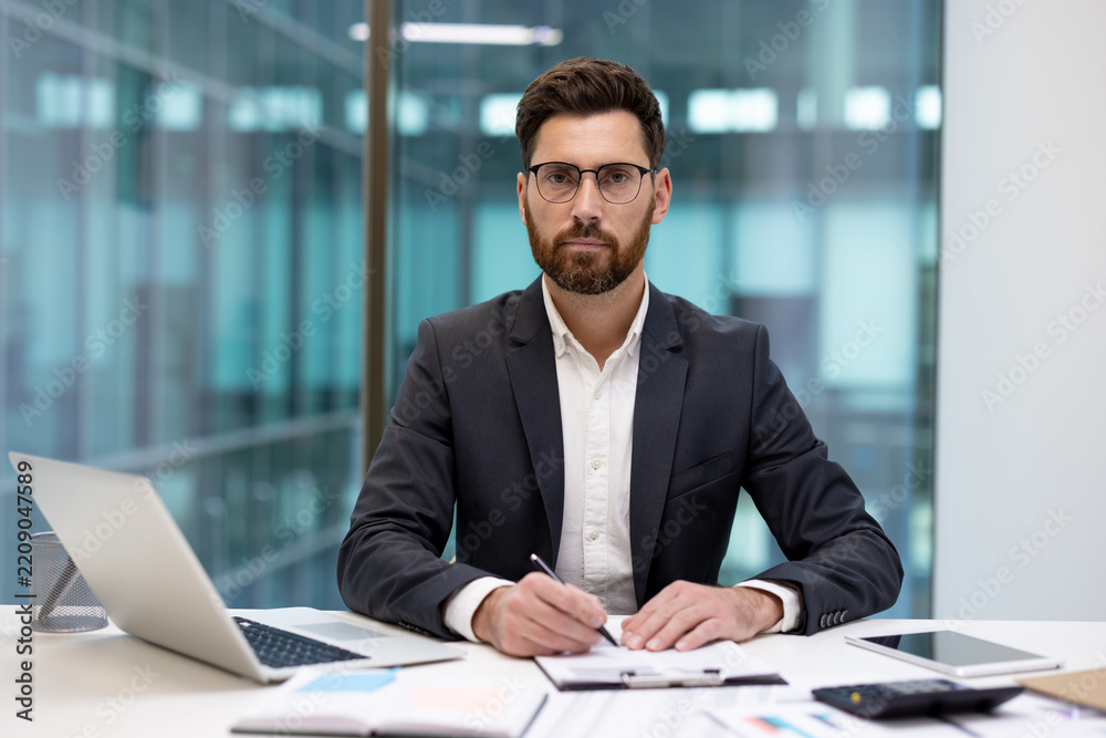 Fototapeta premium A portrait of a serious young businessman in a suit and glasses, sitting at an office desk with a laptop and documents, holding a pen and looking at the camera