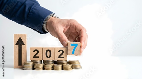 Business professional arranging wooden blocks representing the year 2027 atop stacked currency coins to showcase financial growth projections and investment planning strategies for the coming