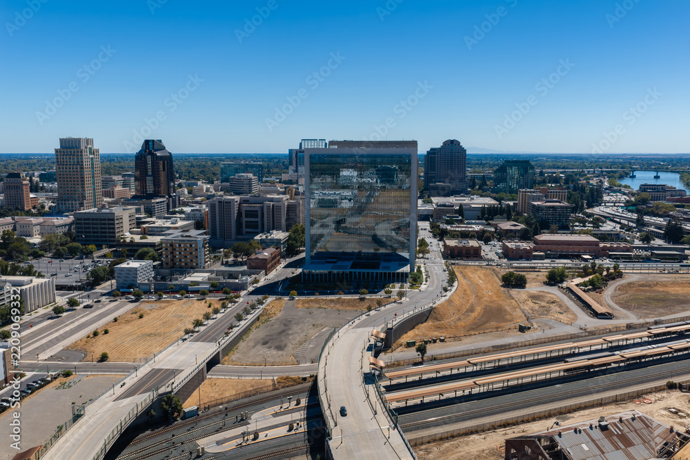 Obraz premium Aerial view shows the downtown Sacramento skyline, featuring modern commercial buildings, railway tracks, highway overpasses, and the Sacramento River in California.