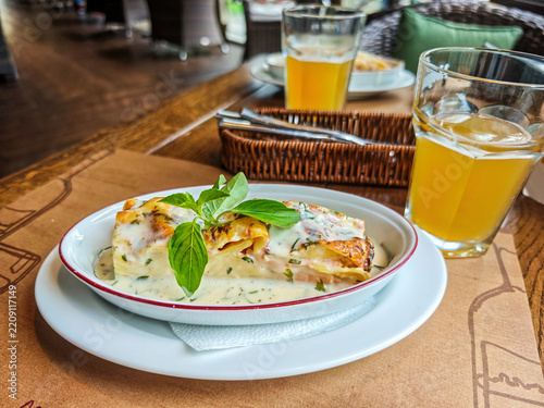 Portion of lasagne or stuffed pasta rolls served with a creamy sauce, garnished with a sprig of basil, along with glasses of drink