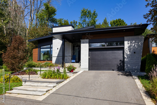 Medium-sized modern house with stone and wood siding, roof overhang and prominent garage door and driveway seen during a sunny fall morning, Quebec City, Quebec, Canada