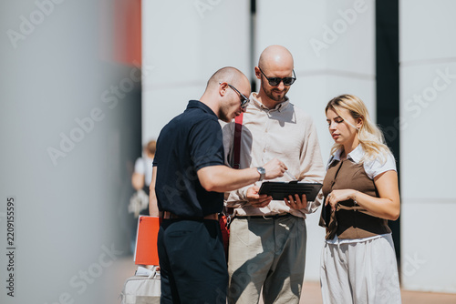 Three business colleagues stand outdoors reviewing a tablet together. They discuss documents and information during a casual outdoor meeting.