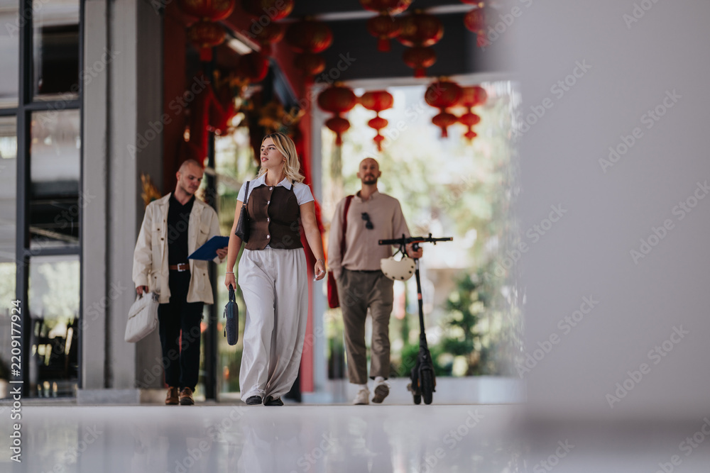 Obraz premium Three friends walk together under red hanging lanterns in a covered urban arcade. A casual group stroll is shown with one person carrying an electric scooter and others holding bags.