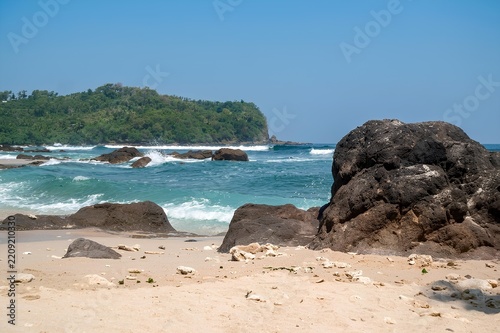Sandy beach with prominent dark rocks in the foreground. Turquoise waves gently crash against the shore.