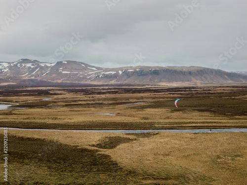 Aerial view of a kitesurfer gliding over a river in a vast Icelandic landscape with snow-capped mountains in the distance in Borgarbyggd, Iceland.
