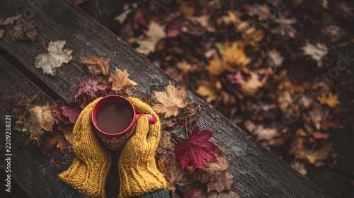 A person wearing yellow knitted gloves holds a coffee mug in their hands, sitting on an old wooden bench surrounded by autumn leaves