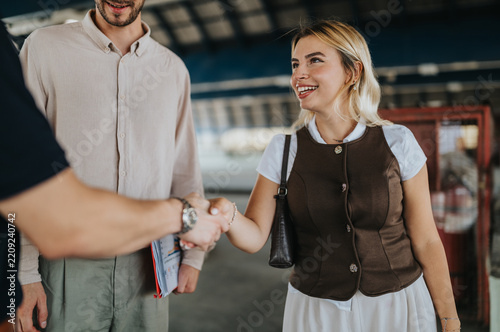 A smiling woman shakes hands with a man while another colleague stands nearby. The friendly introduction takes place on a covered transport platform, showing a casual business meeting.