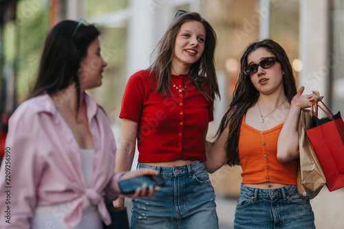 Three young women walk together carrying shopping bags and chatting. Candid street photo of friends enjoying a casual shopping outing in the city.