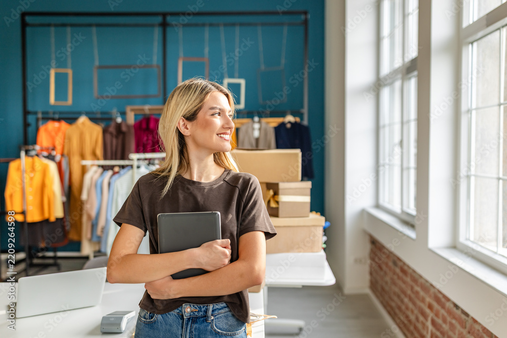 Obraz premium A young woman stands in her clothing business showroom, smiling while holding a tablet. The space features colorful garments displayed on racks, with natural light streaming through large windows, cre