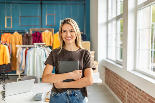 A young woman stands in her small clothing store, smiling while holding a tablet. The shop features colorful garments hanging in the background, with a bright and welcoming atmosphere.