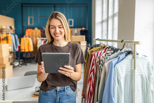 A young woman smiles while using a tablet in her small clothing store. The shop features a variety of colorful garments displayed on a rack, creating a vibrant and inviting atmosphere.