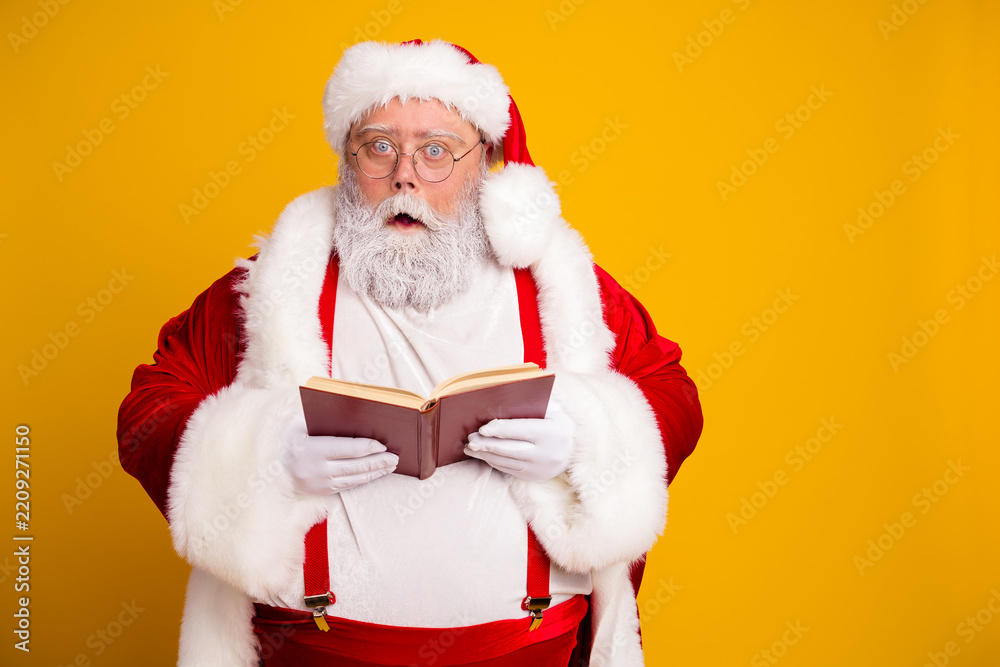 Obraz premium Santa Claus in red suit reading a book with surprised expression on yellow background for Christmas holiday storytelling portrait