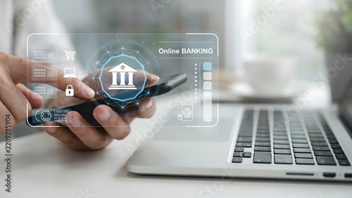 Person's hand using smartphone with holographic online banking interface next to laptop and coffee cup